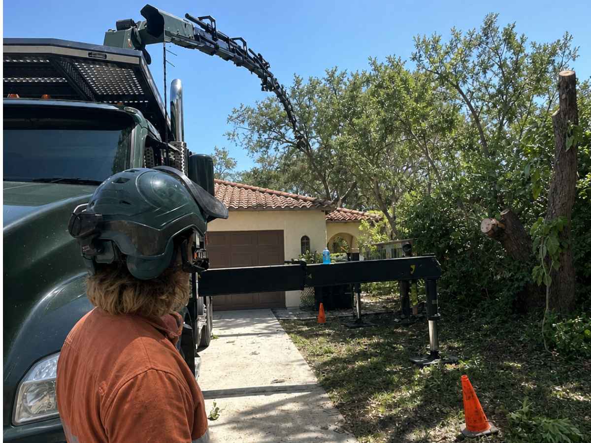 Worker in rust-orange reflective clothing observes a plain green crane truck beside a Spanish Revival bungalow and trimmed trees.