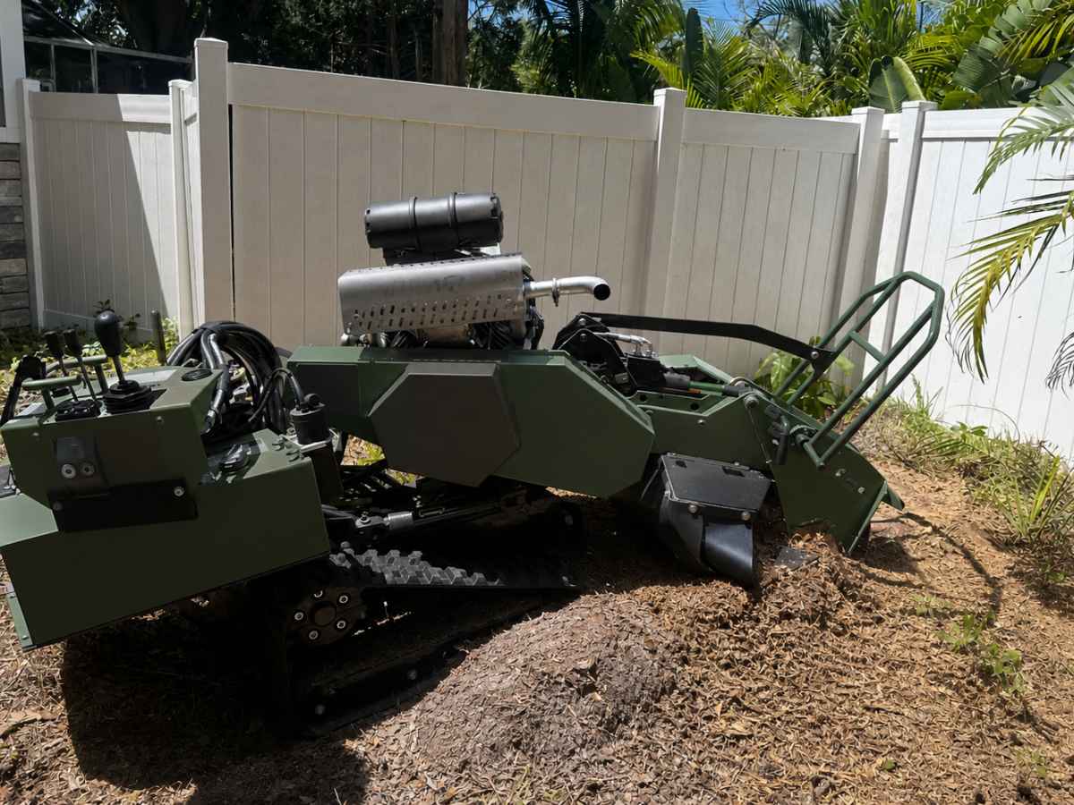 Plain forest-green tracked stump grinder works on a smaller stump beside a gray stone home exterior.