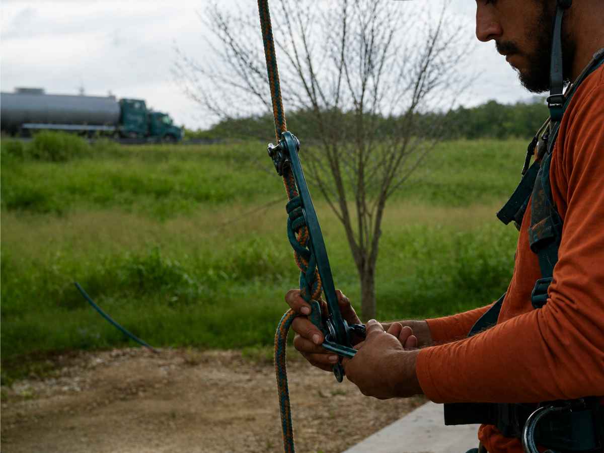 Worker in burnt-orange clothing adjusts plain teal climbing hardware on a rope with a smaller tree and blank tanker beyond.