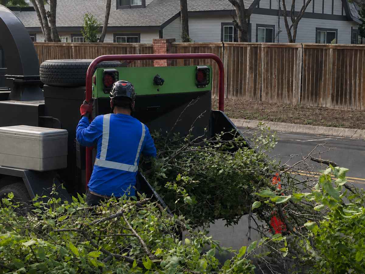 Worker in blue reflective clothing feeds branches into a plain green-and-black chipper near a gray Craftsman home.